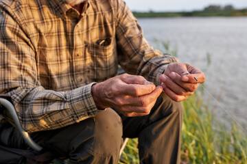 cropped senior male in checkered shirt put the bait of the worm on hook of fishing rod, in nature...