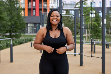 Positive afro american woman in black tracksuit posing at camera outdoors, smiling. Obese...