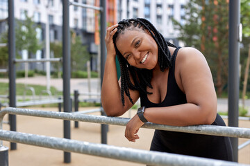 Optimistic african american lady in black sportive outfit taking a break after sport workout. Attractive black obese overweight woman having rest, sport in the morning outdoors. side view portrait