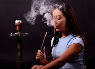 The girl smokes a hookah on a black background. Smoke comes out of the mouth. The pleasure of smoking