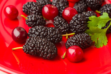 Ripe large black mulberries and red cherries on a red plate, close-up.