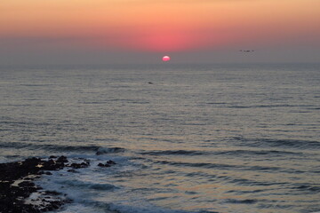 Sunrise over ocean with birds approaching sun, boat on the sea and rocks in the foreground