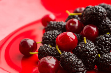 Ripe large black mulberries and red cherries on a red plate, close-up.