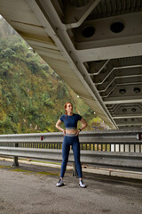 Naklejka premium Attractive caucasian female with red hair having rest taking a break after jogging, at spring day, woman running on fresh air, Slender athelte in blue leggins and top engaged in fitness sport
