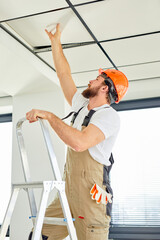 Professional builder on stepladder repairing ceiling, in orange helmet and overalls, side view on...