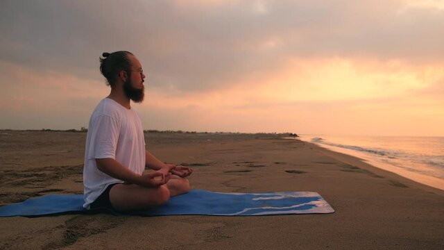 Man practices yoga meditation sitting in padmasana during sunrise. Man sits in lotus position at beach opposite rising sun and its reflection in sea
