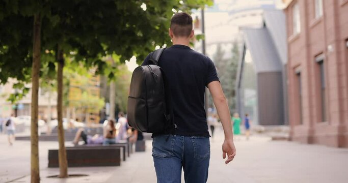 Rear view man walking in city with black leather backpack on his shoulders, slow motion