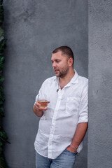 Close-up portrait of a young stylish man holding a glass of white wine, drinking. A guy on a date is wearing a white shirt, blue jeans, standing against a gray wall. The groom laughs at the wedding