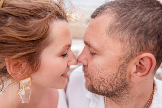 A very close-up portrait of a couple in love who looks at each other, touching nose to nose. The lovers in white laugh. Young woman with natural natural makeup, glass earrings. A man with a beard