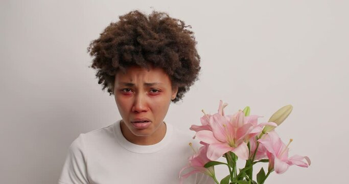 Sick woman with curly hair suffers from allergic rhinitis sneezes and has swelling eyes has allergy to lilies wears white t shirt poses indoor. Reaction to trigger. Hypersensitive female feels unwell
