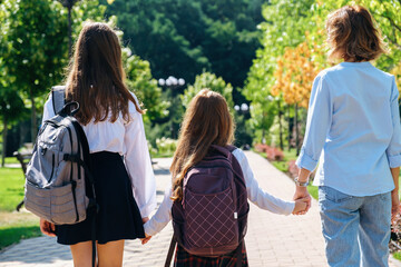 Two daughters in school uniform walking together with mother to school down the street, holding...