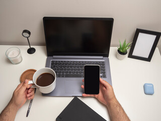 Man's hands on a white table with a laptop typing on the keyboard next to a mobile phone a black notebook a cup of coffee a clock a white candle a small bush and a blank photo frame
