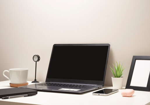 Picture of laptop on a white table with a mug a clock a small bush a notebook a phone a picture frame and a headphone case