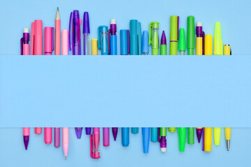 Rainbow stationery collection of pens, ballpoints and pencils on a light blue background with copy space. Lots of colorful office and school supplies in a row under a paper sheet.