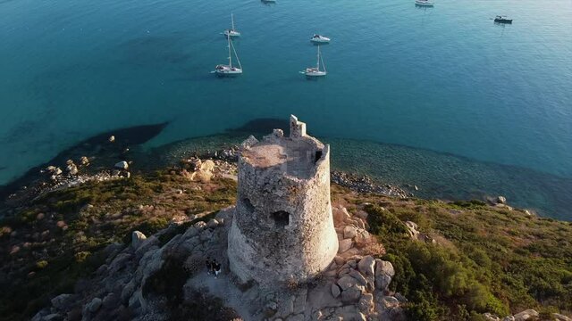 Birds eye view of Torre di Porto Giunco tower standing tall on island surrounded by sea with yachts sailing and people exploring and taking pictures in Italy