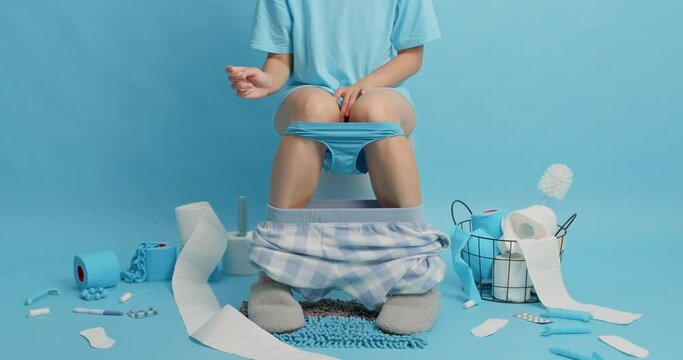Unknown woman sits on toilet bowl leans to floor takes sanitary napkin has menstuation wears comfortable pajama pants pulled down on legs surrounded by toilet paper rollers against blue background
