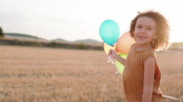 Track shot of happy boy running over countryside field and holding balloons