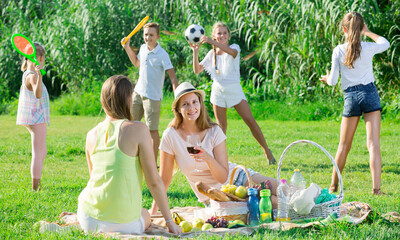 Fototapeta premium Two cheerful mums enjoying picnic outdoors, while kids playing active games