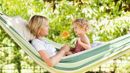smiling mom and little girl daughter child blue eyes with blond curly hair, together lying on the hammock in the green home garden, plays with lily flower, happy family and spring time concept