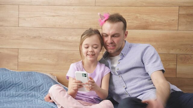 Cute child girl showing something funny on smartphone to dad, they sit together, enjoying free time at weekends, holidays. Cosmetics and accessories on bed.