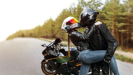 Motorcyclist in leather jacket and helmet stands next to sports motorcycle and hugged girl in helmet and tilted her, blurred background, copy space