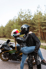 Motorcyclist in leather jacket and helmet stands next to sports motorcycle and hugged girl in helmet and tilted her, blurred background, copy space