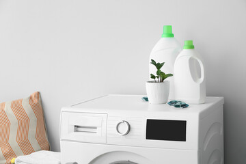 Modern washing machine with detergent and houseplant near light wall