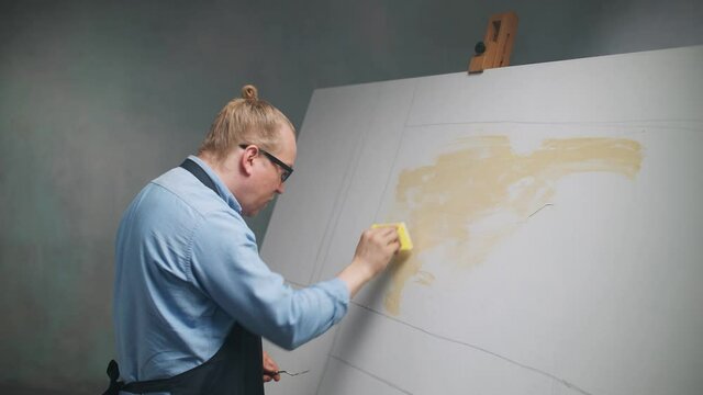 Slow motion, creative person at work, a talented adult man artist covers the canvas with beige oil paint, priming and draws the background.