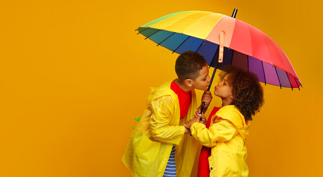 kissing Black childs girl and a boy with yellow raincoat under a colored umbrella on colored yellow background.