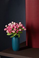 Subject shot of graceful bouquet of tender pink tulips with ornamental herbs. Sea-green vase with bunch of flowers is located on the brown table near red curtain against gray wall. 