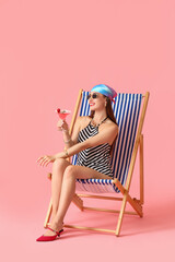 Beautiful young woman with cocktail sitting on deck chair against color background