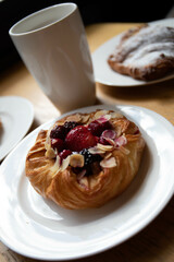 an almond croissant and a bun with strawberry slices lie on a white plate next to a white cup of coffee cappuccino and latte food fresh pastries bakery handmade kitchen cafe breakfast health