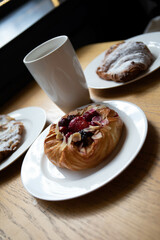 an almond croissant and a bun with strawberry slices lie on a white plate next to a white cup of coffee cappuccino and latte food fresh pastries bakery handmade kitchen cafe breakfast health