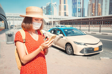 Female tourist in a medical protective mask during the Covid pandemic calls a taxi using an...