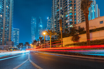 A photo with a long exposure with visible traces from the headlights of passing cars and illuminated skyscrapers in the Dubai Marina district