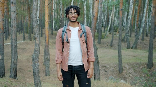 Slow motion portrait of joyful mixed race guy tourst standing in green woods with backpack and looking at camera with carefree smile. Youth and tourism concept.
