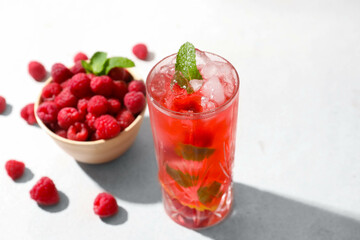 Glass of delicious lemonade and bowl with raspberry on light background