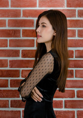 Side way portrait of young beautiful attractive Asian female model in black revealing dress smiling and standing against orange brick wall background