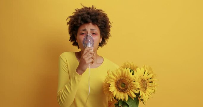 Difficult to beath. Unhappy curly African American woman suffers from suffocation shortness of breathing asthmatic fit wears oxygen mask allergic to sunflowers poses against yellow background