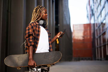 Obraz premium Portrait of modern african girl with skateboard. Young stylish woman using the phone.