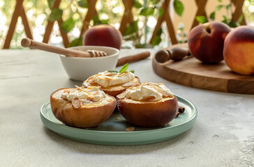 Selective focus of baked peaches with cinnamon, almond petals, cream cheese, and honey on a light concrete background. Fruits on a plate with mint. Summer garden snack. 