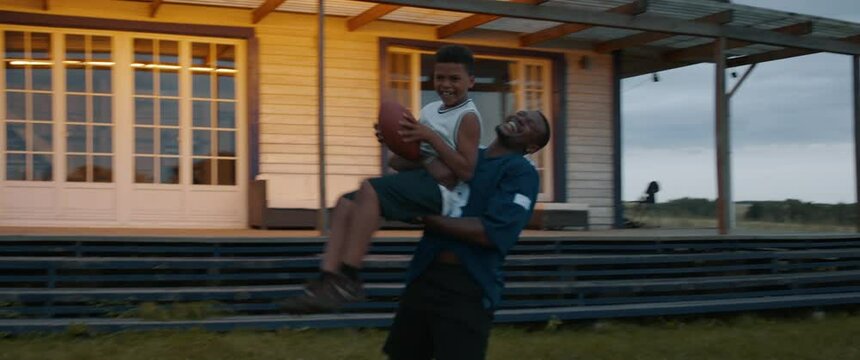 TRACKING African American father and son playing football together on a lawn in front of their house in the evening. Shot with 2x anamorphic lens