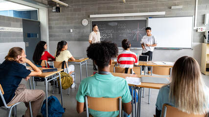 Teen asian boy high school student giving a presentation in class to his multiracial classmates and teacher. Horizontal banner image.