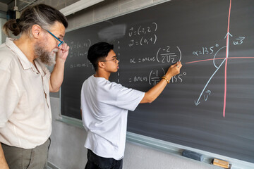 Caucasian mature man teacher supervises teen asian high school boy student writing maths exercise...