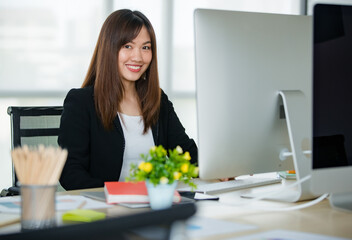 Young attractive Asian woman in black business suit sitting down working on computer desktop in modern looking office. Concept for modern office lifestyle