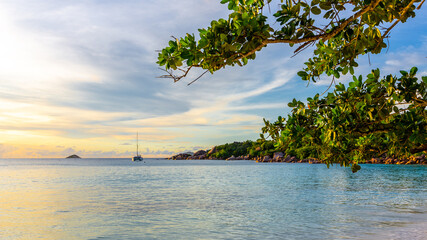 Obraz na płótnie Canvas Idyllic sunset on Anse Lazio beach with catamaran anchored in the distance, calm turquoise water and tropical trees and leaves around.