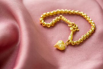 The gold bracelet has a heart pendant placed on a pink gold fabric.