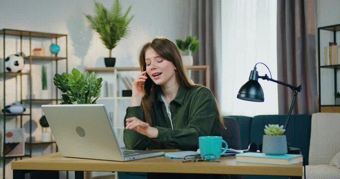 Attractive smiling happy young smart girl working on computer in home office and talking on mobile simultaneously drinking tea,front view,slow motion
