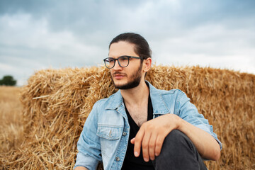 Naklejka na ściany i meble Portrait of young man sitting near haystacks on background of clouds.