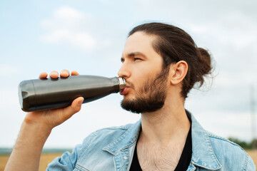 Portrait of guy with ponytail hairstyle drinking water from thermo bottle. - obrazy, fototapety, plakaty